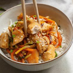 A bowl of stir-fried chicken and vegetables over rice, garnished with sesame seeds.