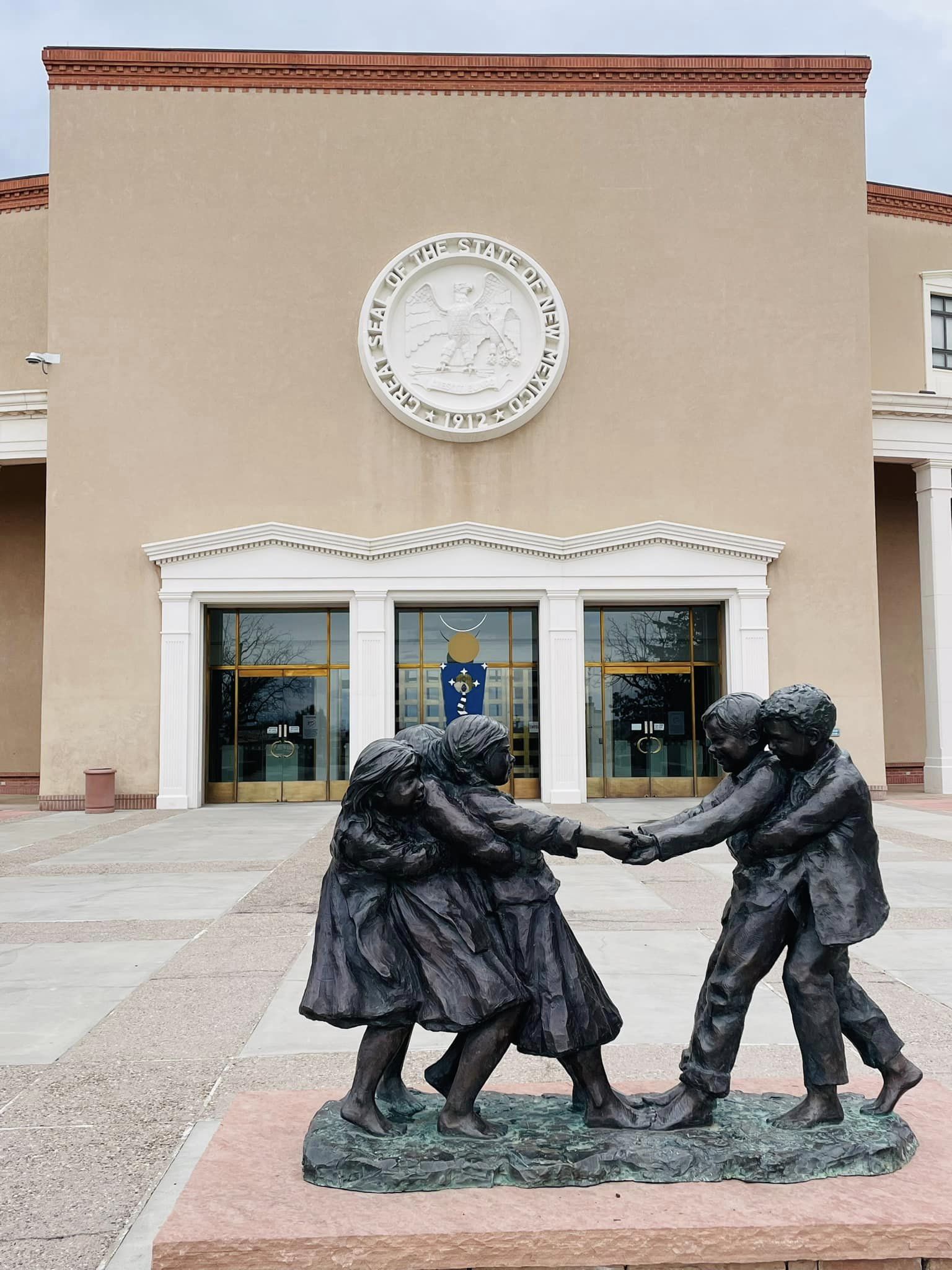 Bronze sculpture of children playing in a ring-around-the-rosy in front of a building with the New Mexico state sea.