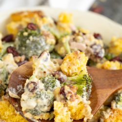 A wooden spoon scooping broccoli and cauliflower salad with cranberries and creamy dressing from a serving bowl.