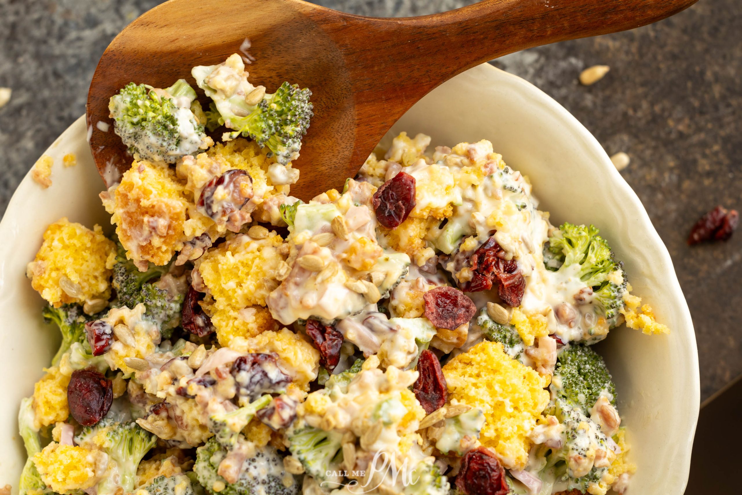 A bowl of broccoli and cauliflower salad with cranberries and nuts, mixed with a creamy dressing, and a wooden serving spoon.