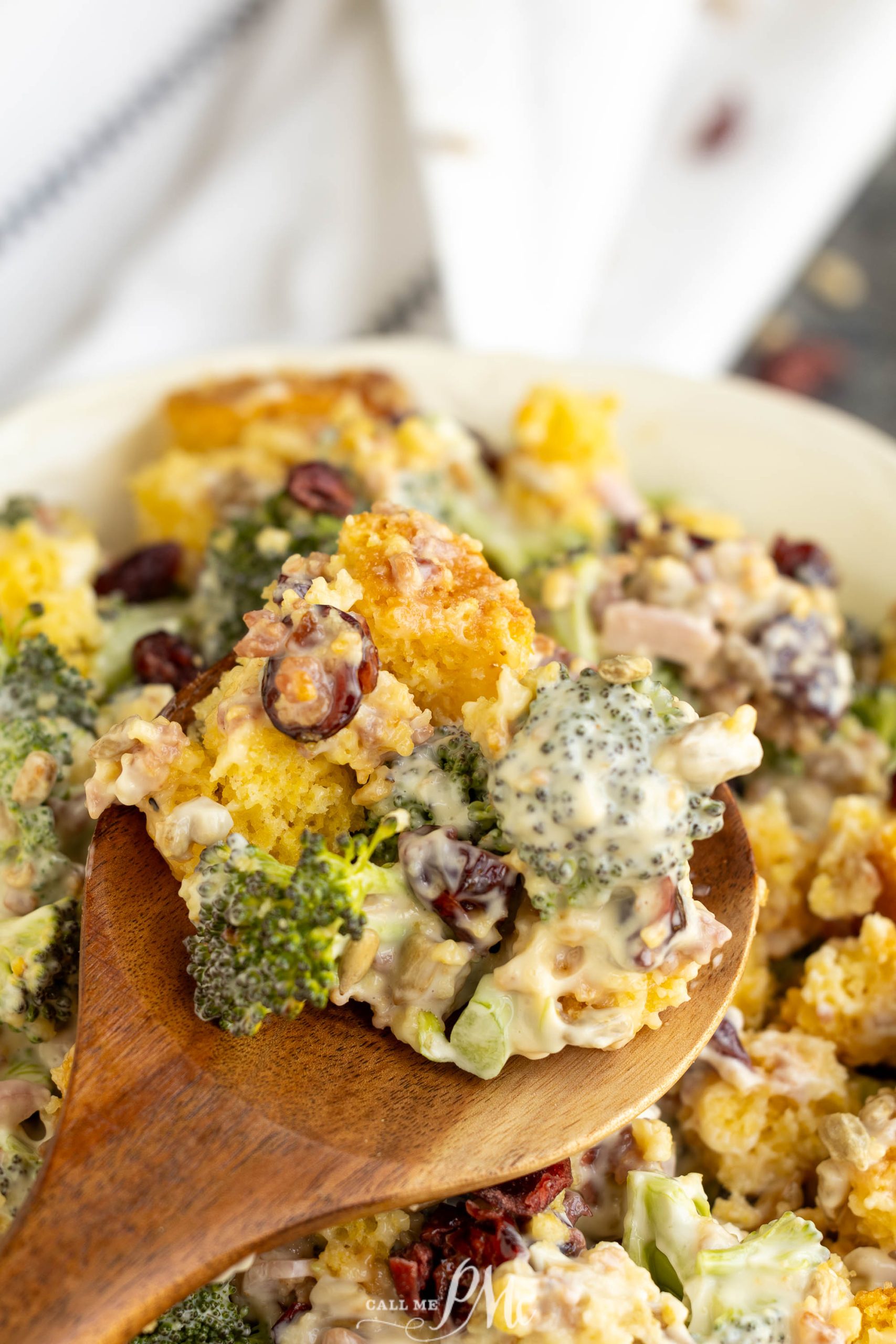 A bowl of broccoli salad with raisins, nuts, and a creamy dressing, served with a wooden spoon.