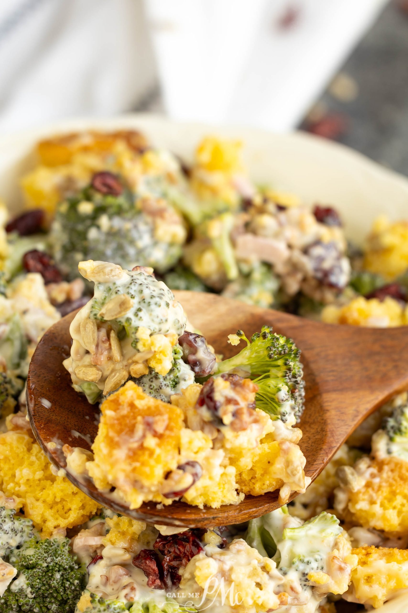 A wooden spoon scooping a colorful broccoli salad with cornbread, cranberries, and a creamy dressing from a white dish.