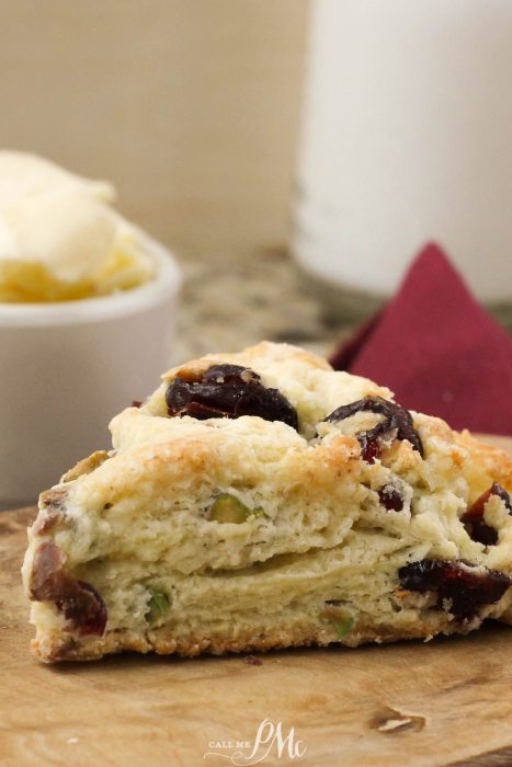 Close-up of a cranberry and pistachio scone on a wooden table, accompanied by a scoop of butter and a glass of milk in the background.