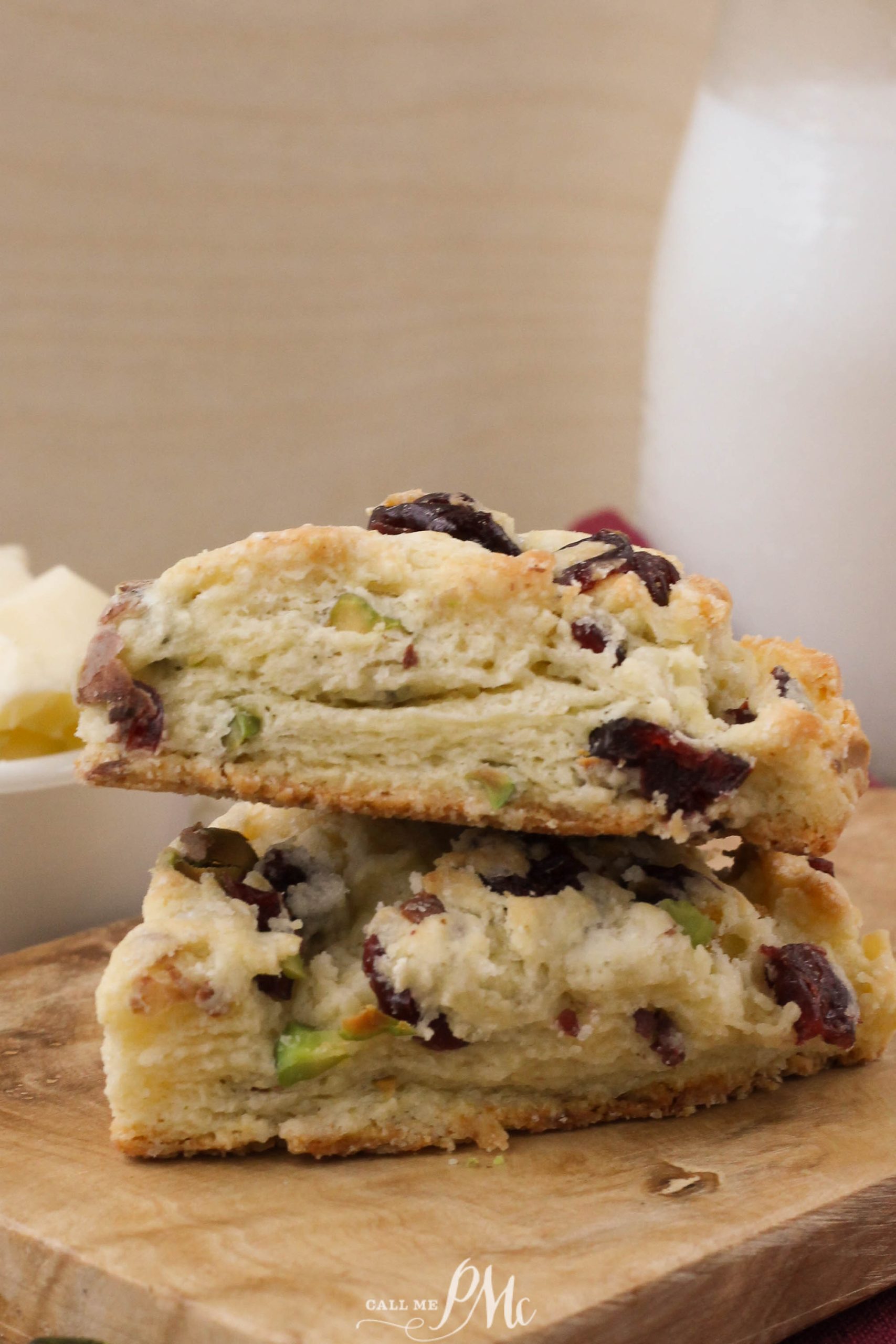 A close-up of cranberry and pistachio scones stacked on a wooden board, with butter and a glass of milk in the background.