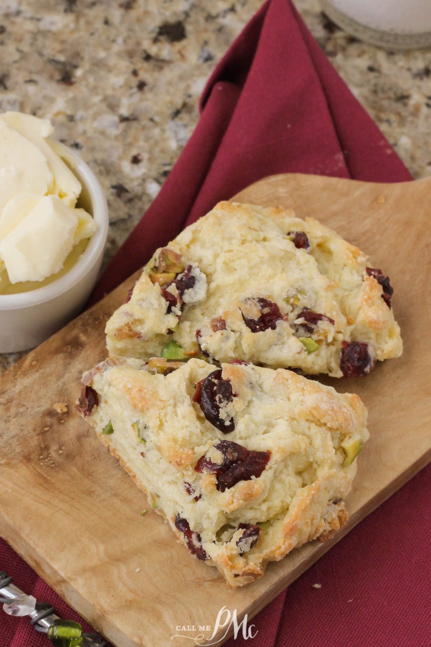 Cranberry and pistachio scones on a wooden board with a bowl of butter, next to a red napkin.