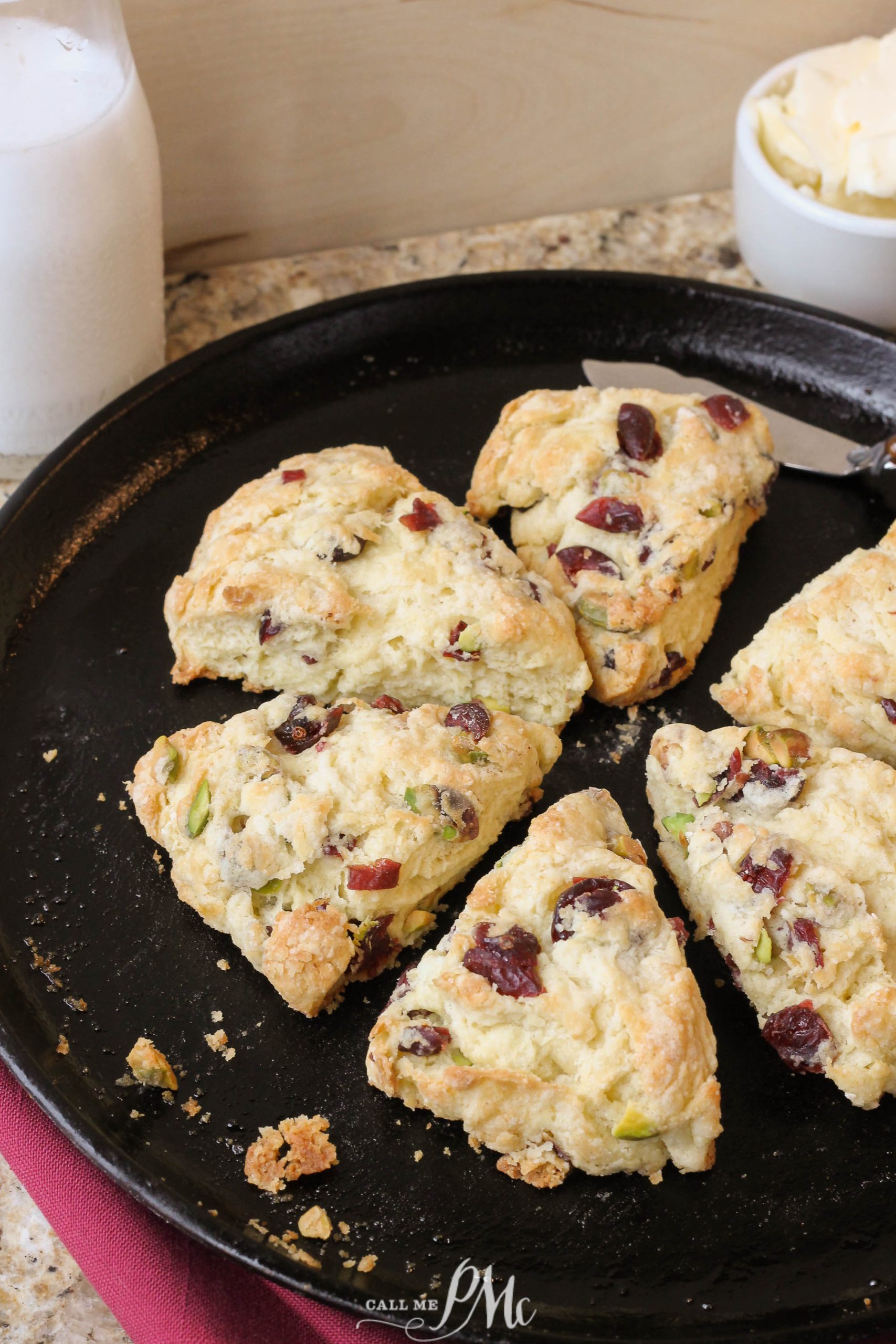 A platter of homemade cranberry and pistachio scones on a black round dish, with a bottle of milk and a bowl of butter in the background.