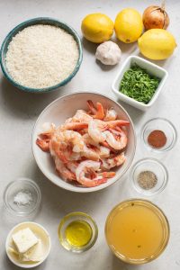 Ingredients for a seafood dish displayed on a countertop, including shrimp, rice, lemons, garlic, chopped herbs, and various seasonings.