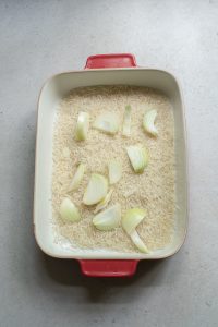 Uncooked rice in a red ceramic baking dish with chunks of raw onion scattered on top, prepared for cooking.