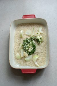 Uncooked rice in a red baking dish with chopped fennel and herbs on top, ready for cooking.