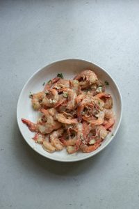 A bowl of cooked shrimp garnished with herbs on a gray surface.