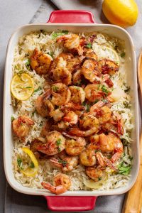 A dish of roasted shrimp over seasoned rice in a ceramic baking dish, garnished with lemon slices and parsley.