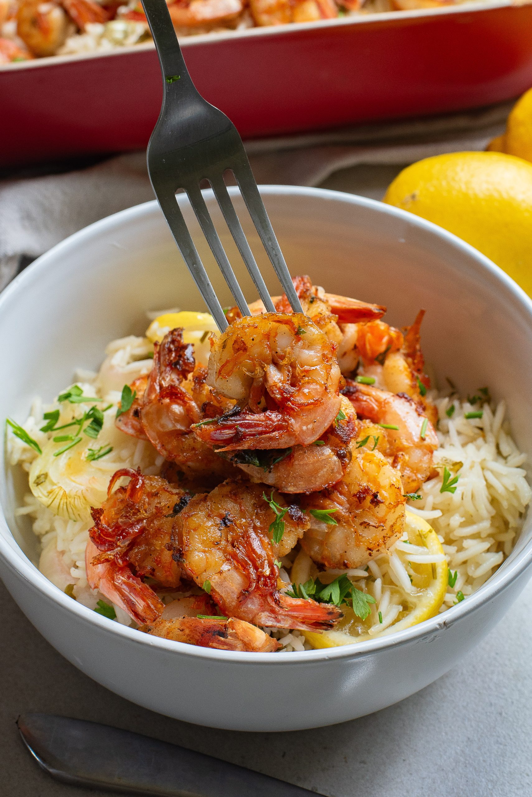 A bowl of cooked shrimp served over rice with lemon wedges on the side, a fork piercing some shrimp.