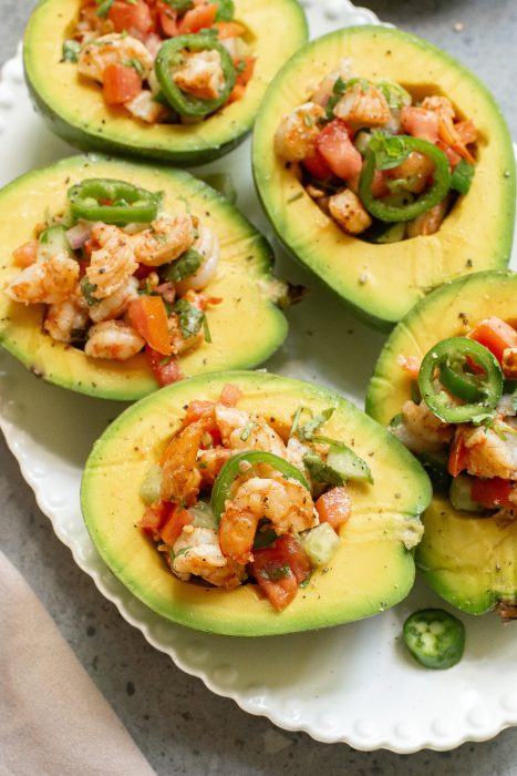 Halved avocados filled with shrimp and diced vegetables, garnished with jalapeño slices, served on a white plate.