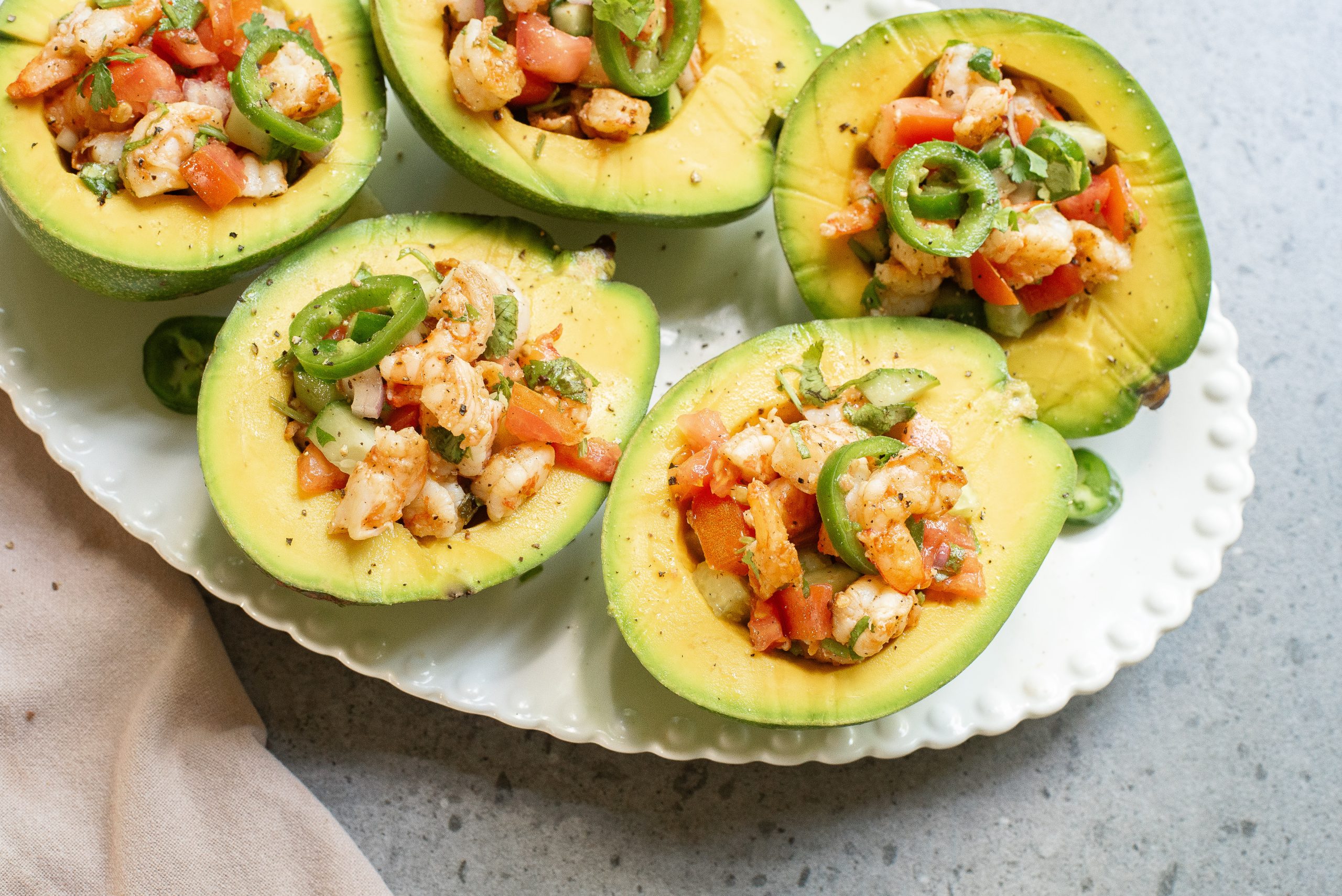 Halved avocados filled with shrimp and diced vegetables, garnished with jalapeño slices, served on a white plate.