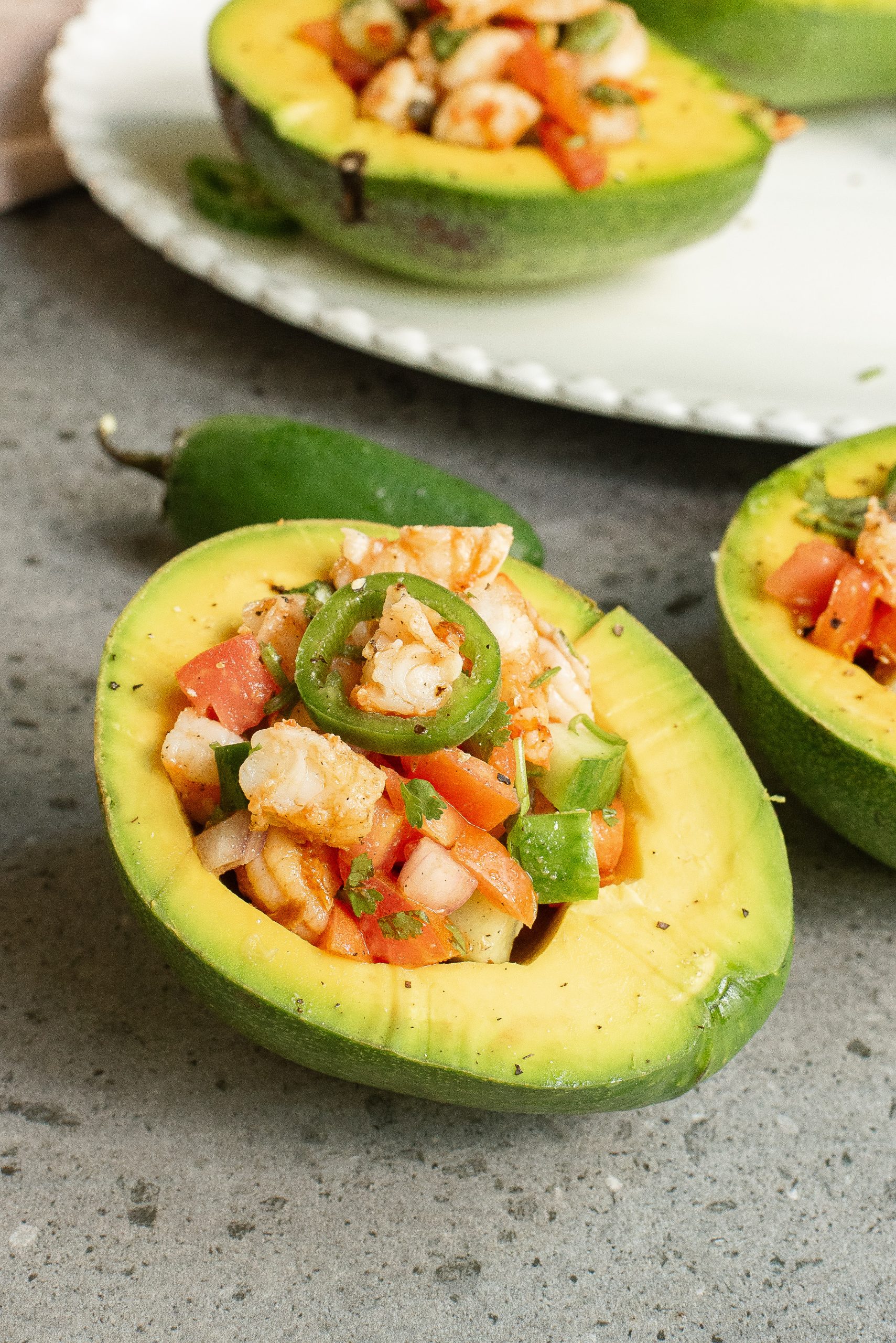 A halved avocado filled with a shrimp salad, topped with a slice of jalapeño, on a white plate.