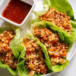 Lettuce wraps filled with savory minced meat and grains, served with a side of red sauce on a white plate.