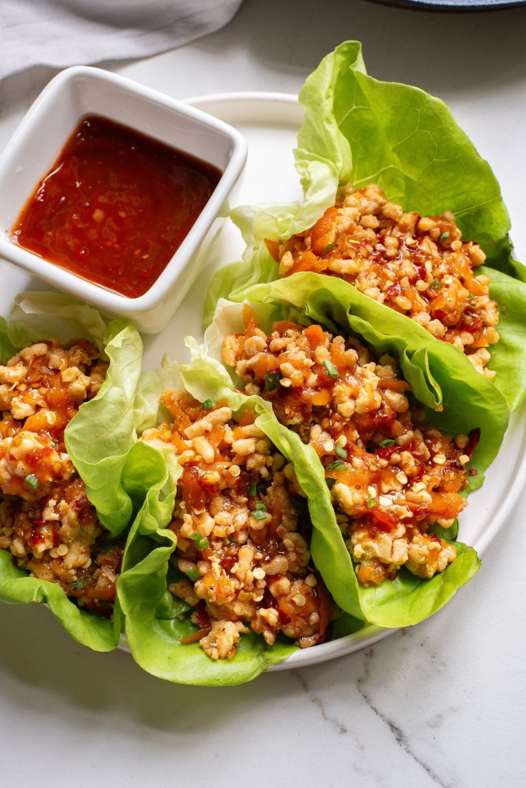 Lettuce wraps filled with savory minced meat and grains, served with a side of red sauce on a white plate.