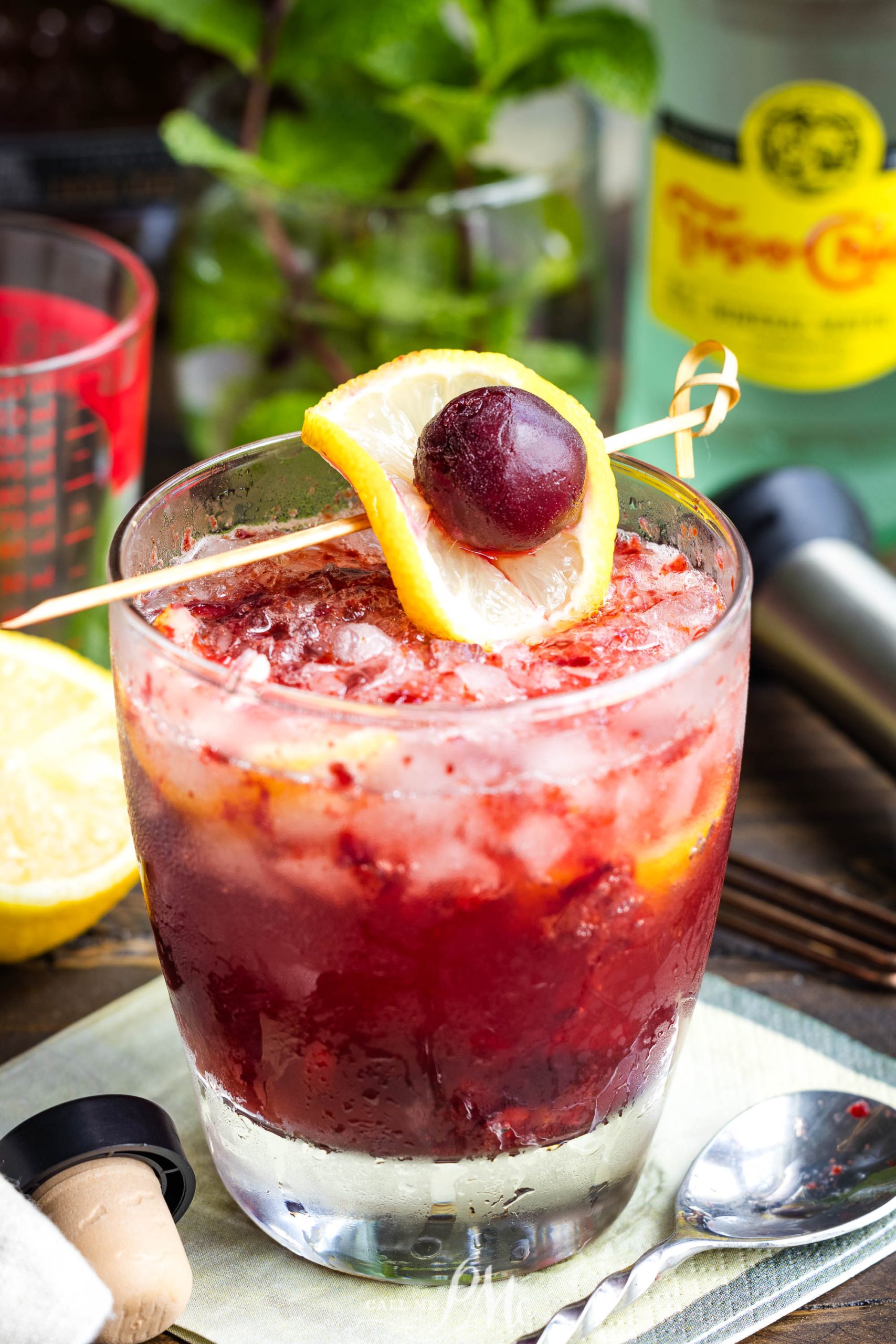 Iced cocktail in a glass garnished with a lemon slice and a cherry, with a bottle and bar tools in the background.