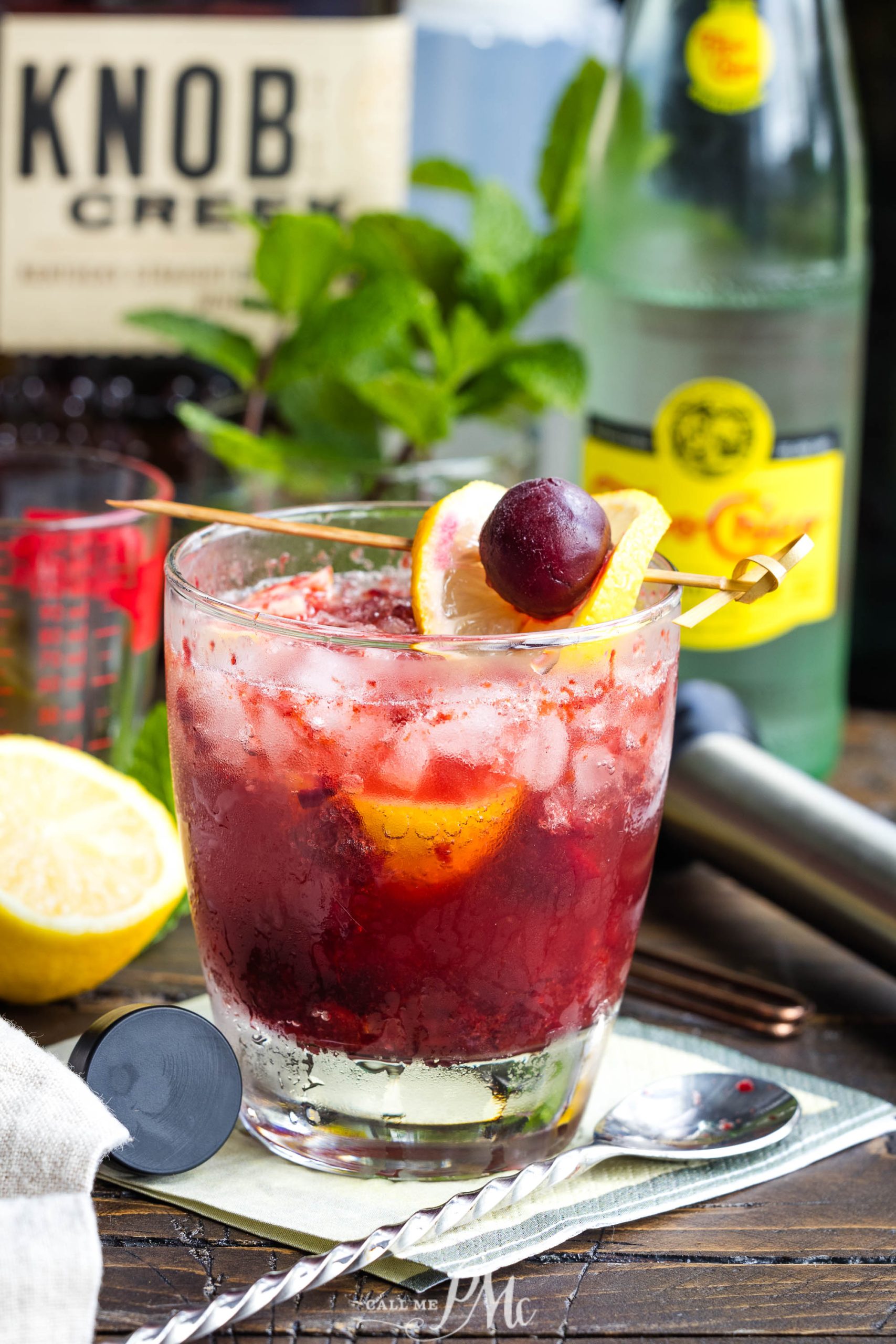 A refreshing cocktail with crushed ice, lemon slices, and a cherry, garnished with mint, served on a wooden table next to a bottle of tonic water and spirits.