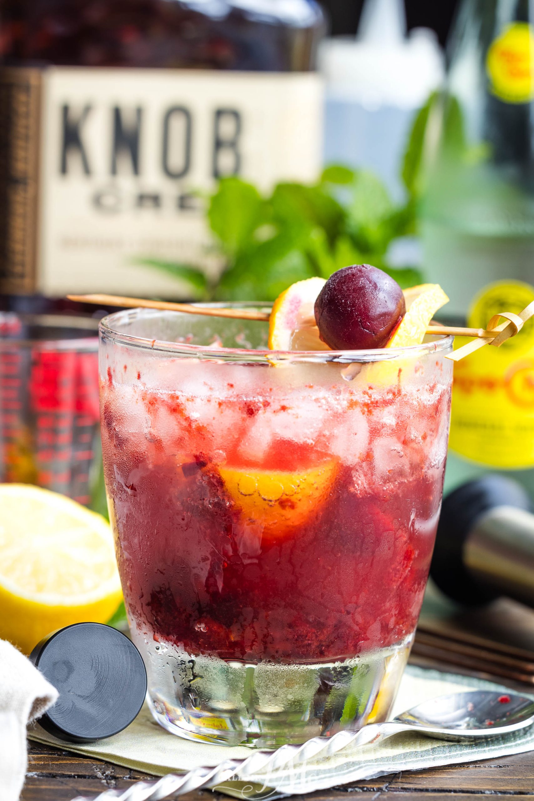 A close-up of a vibrant red cocktail, Fizzy Cherry Bourbon Sour, with crushed ice, garnished with a lemon slice and cherry, in a glass tumbler, with a blurred bottle labeled "knob creek" in the background.