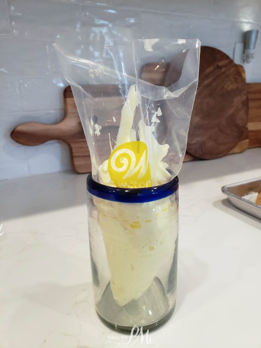 A jar of cream with a wilton brand label seen through a ripped plastic bag on a kitchen counter.