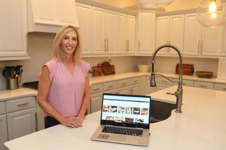A woman in a pink blouse stands in a kitchen with white cabinets, smiling beside an open laptop displaying a webpage.