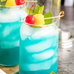 Two glasses of blue cocktails with ice, garnished with mint leaves, a maraschino cherry, and a pineapple chunk, are placed on a wooden board.