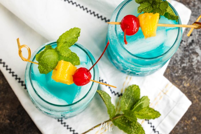 Two Blue Hawaiian Cocktailwith ice, garnished with skewers of a red cherry, yellow pineapple chunk, and fresh mint, placed on a white cloth with black stitching. Mint leaves are scattered nearby.