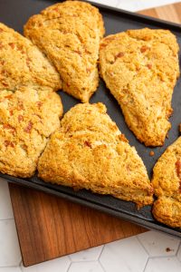 A tray of six golden-brown scones, arranged in neat, spaced intervals on a black baking sheet, rests on a wooden cutting board.
