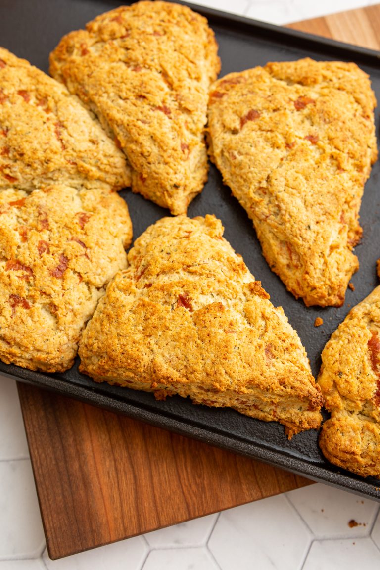 A tray of six golden-brown scones, arranged in neat, spaced intervals on a black baking sheet, rests on a wooden cutting board.