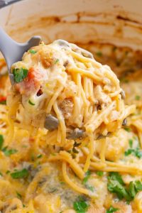 A ladle scoops a portion of cheesy baked spaghetti with tomatoes and herbs from a pot.