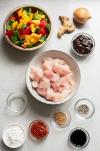 Ingredients for a recipe laid out on a counter, including chopped vegetables, diced chicken, ginger, onion, and various spices and sauces.