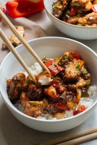 Bowl of stir-fried chicken with vegetables and rice, picked up with chopsticks, on a textured table with ingredients in background.