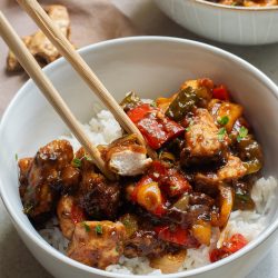 Bowl of stir-fried chicken with vegetables and rice, picked up with chopsticks, on a textured table with ingredients in background.