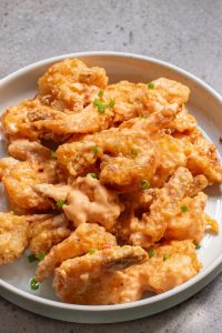 A white plate filled with crispy fried shrimp covered in a creamy, orange sauce, garnished with chopped green onions.