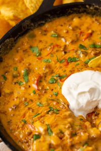A skillet filled with cheesy nacho dip topped with chopped herbs and a dollop of sour cream, with a few tortilla chips visible at the edge.