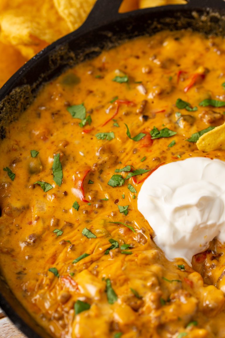 A skillet filled with cheesy nacho dip topped with chopped herbs and a dollop of sour cream, with a few tortilla chips visible at the edge.