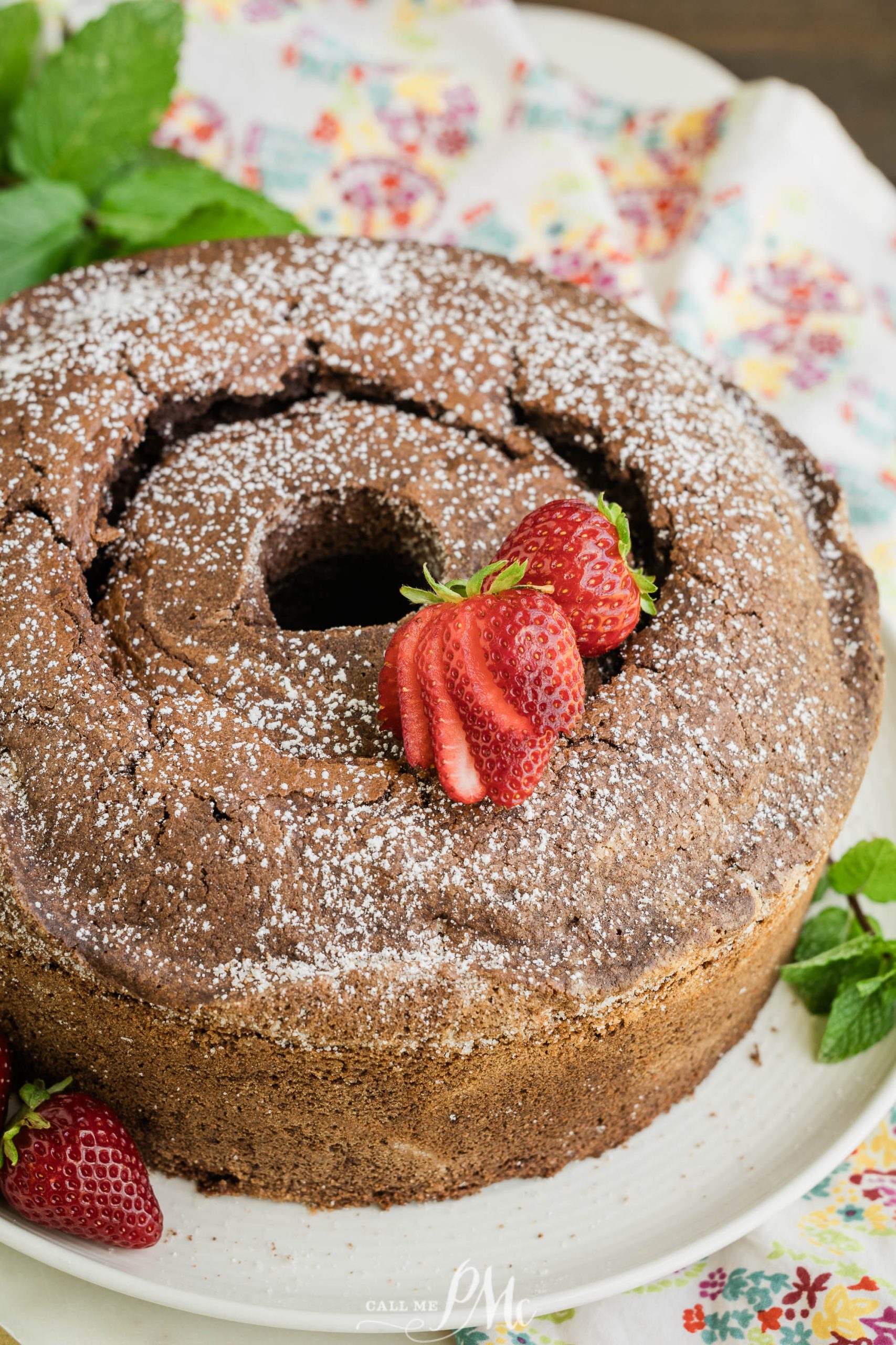 A chocolate bundt cake sprinkled with powdered sugar and garnished with fresh strawberries on a floral plate.