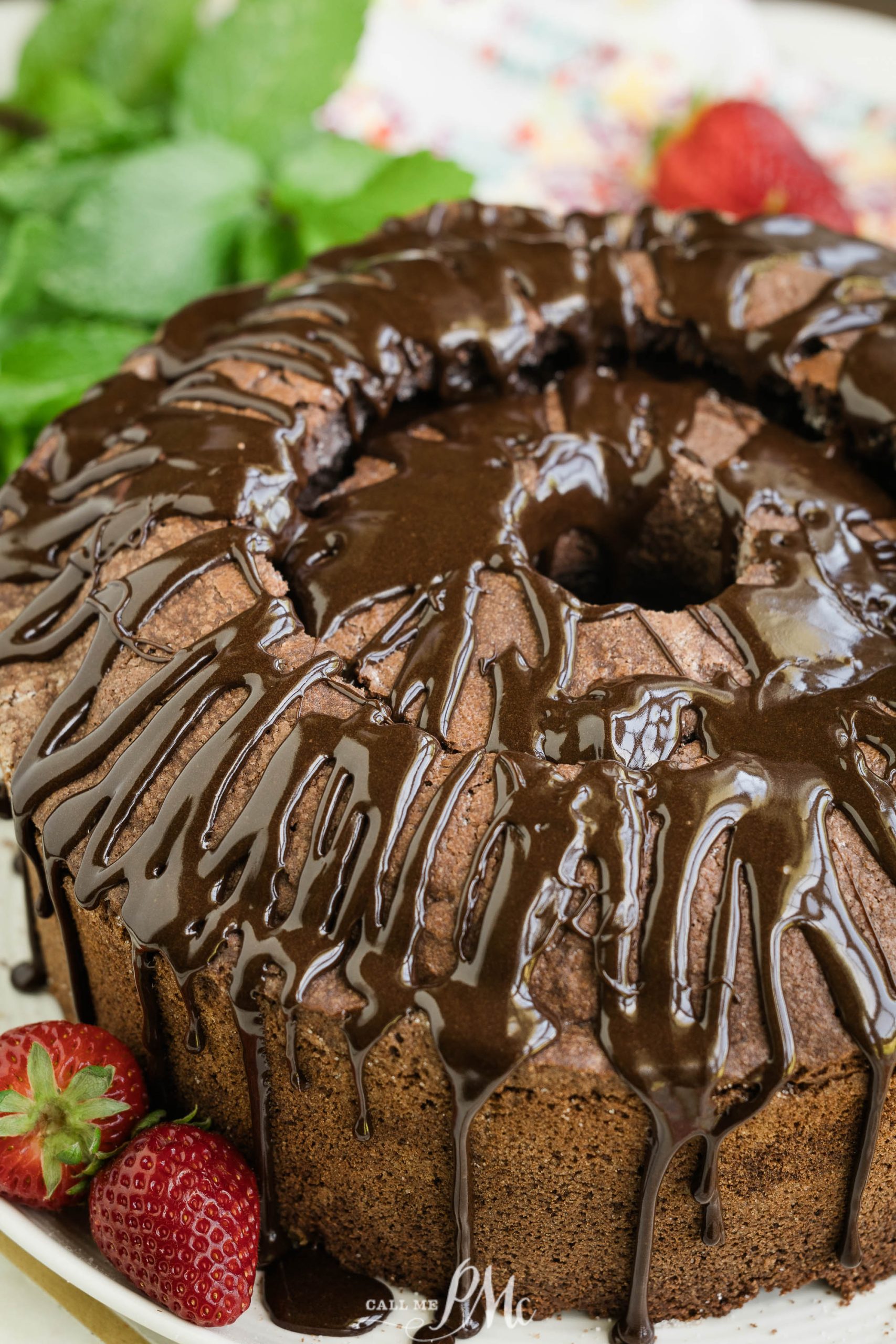 A bundt cake with a rich chocolate glaze drizzling down its sides, garnished with a fresh strawberry, presented on a plate with mint leaves in the background.