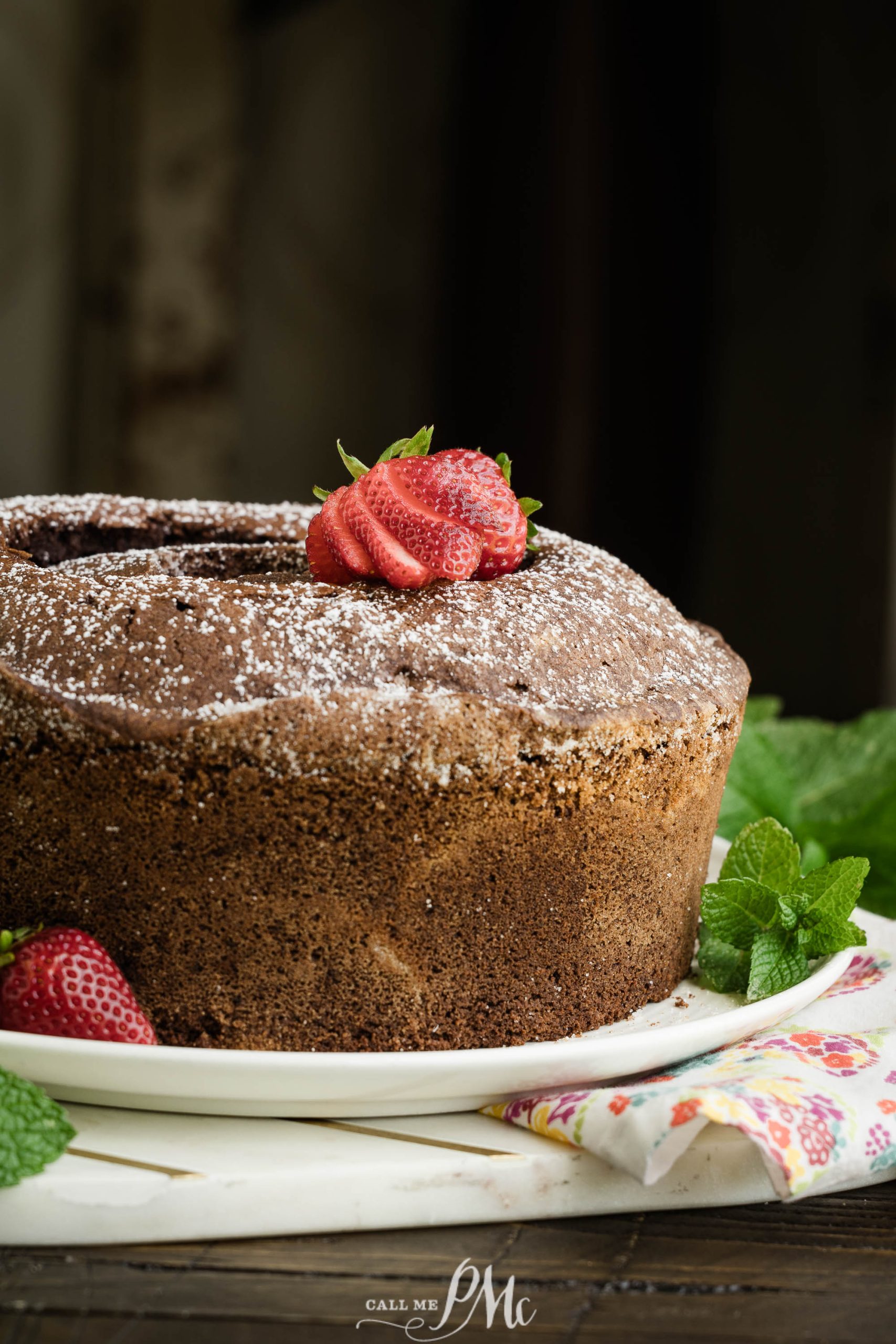 A bundt cake sprinkled with powdered sugar, garnished with fresh strawberries and mint leaves on a white plate.