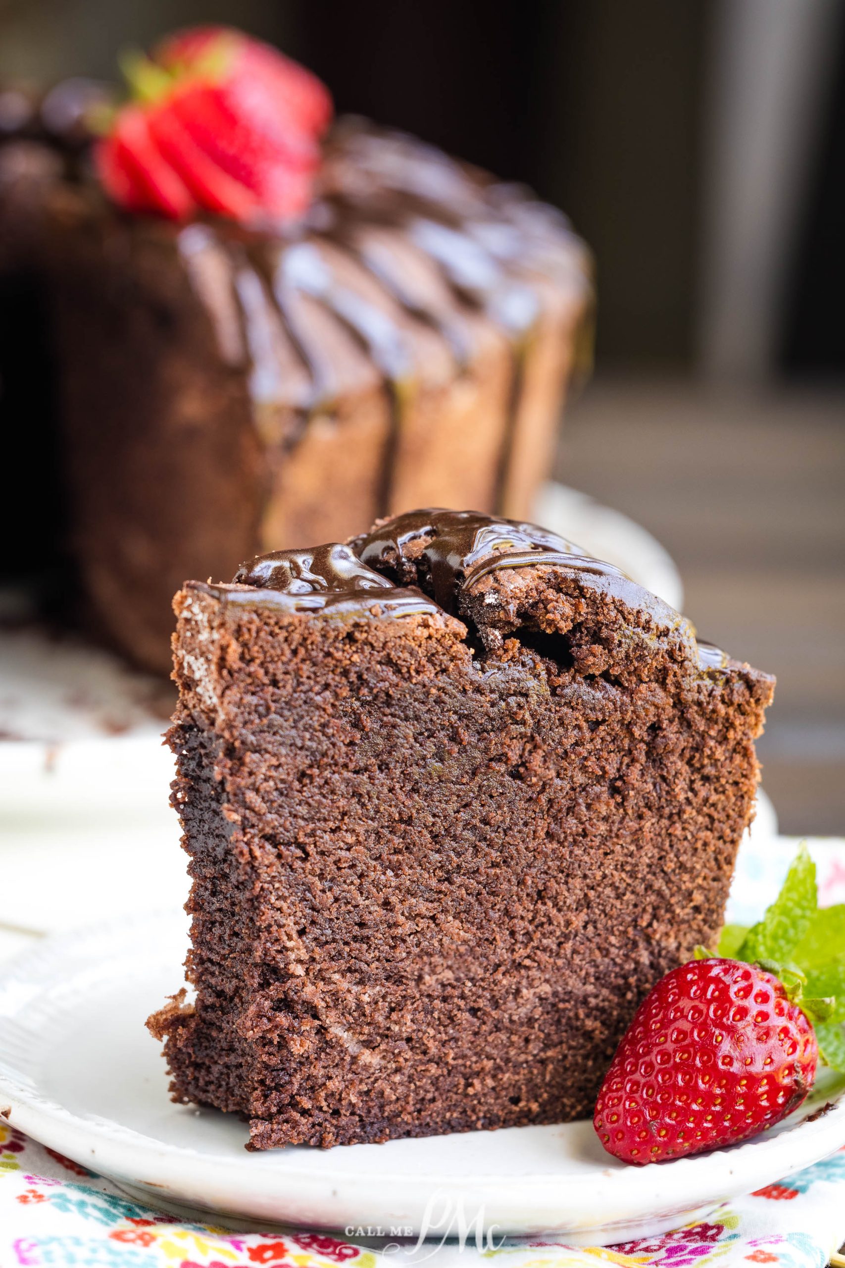 A slice of dessert topped with glossy chocolate ganache, garnished with a fresh strawberry, served on a decorative plate.