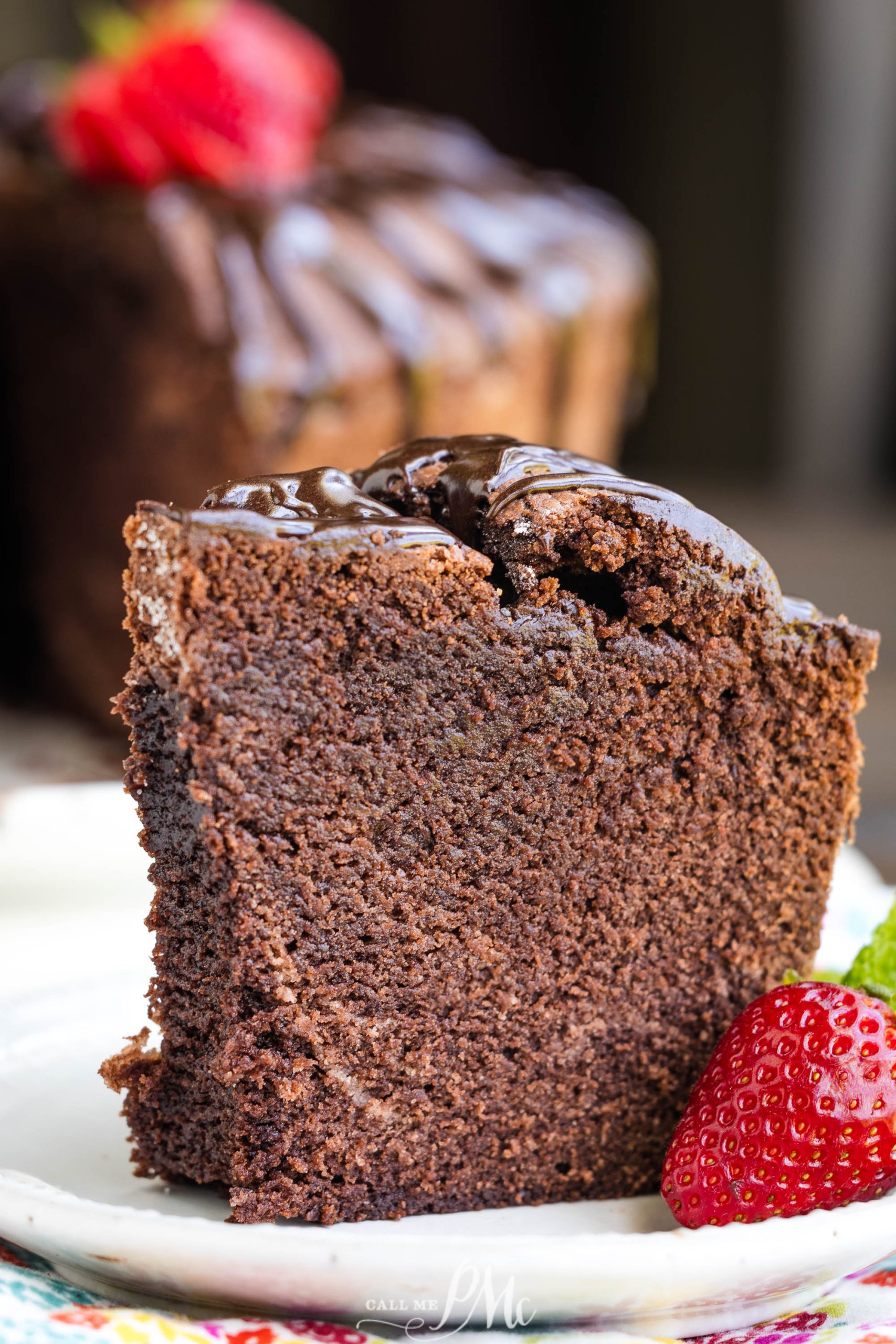 A slice of rich chocolate cake with glossy chocolate icing, served on a white plate, garnished with a fresh strawberry on the side.