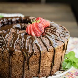 A chocolate bundt cake topped with a glossy chocolate glaze and garnished with fresh strawberries and mint leaves on a floral tablecloth.