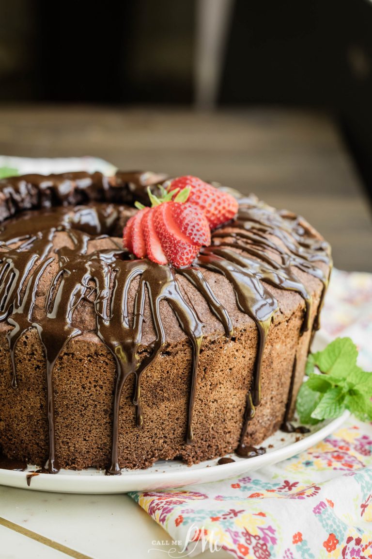 A chocolate bundt cake topped with a glossy chocolate glaze and garnished with fresh strawberries and mint leaves on a floral tablecloth.