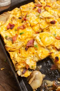 A close-up of a fresh-baked, sliced potato and cheese casserole on a baking tray, topped with bits of bacon and garnished with herbs. A portion has been served, revealing the layers inside.
