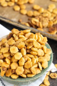 A close-up of a bowl filled with hexagon-shaped, seasoned snack crackers. More crackers can be seen scattered on a baking sheet in the background.