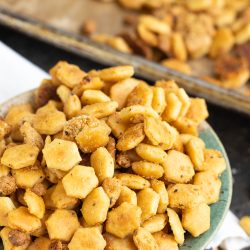 A close-up of a bowl filled with hexagon-shaped, seasoned snack crackers. More crackers can be seen scattered on a baking sheet in the background.
