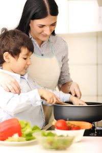 A woman and a young boy cooking together in a kitchen, smiling as they stir a pot on the stove with assorted vegetables around them.