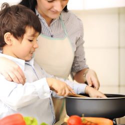 A woman and a young boy cooking together in a kitchen, smiling as they stir a pot on the stove with assorted vegetables around them.