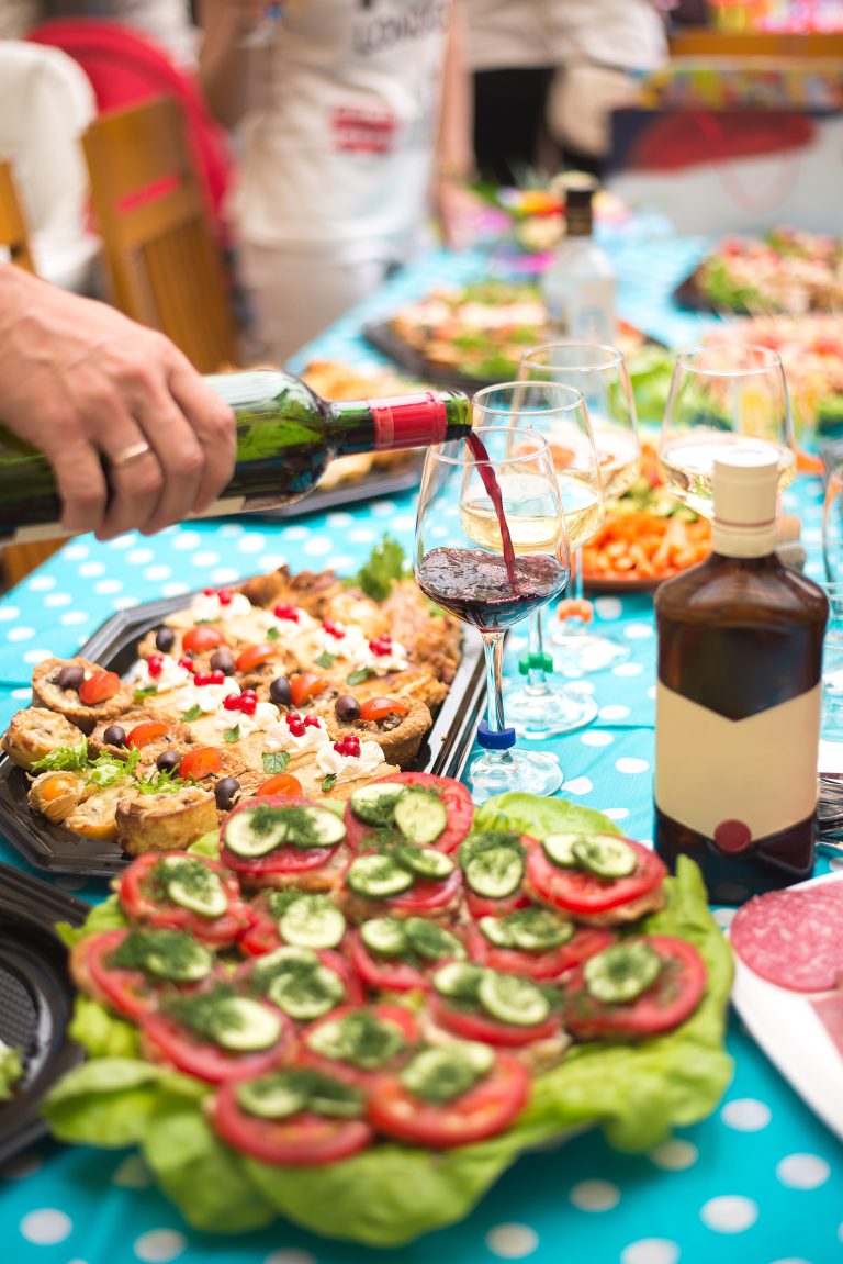 A person pours red wine into a glass at an outdoor dining table filled with colorful dishes of food, including a platter of bruschetta.